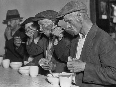 Four men wearing hats and coats eat soup at a counter, each holding a spoon and a piece of bread. The setting appears to be a simple dining area or soup kitchen. The image is in black and white, suggesting it is historical.